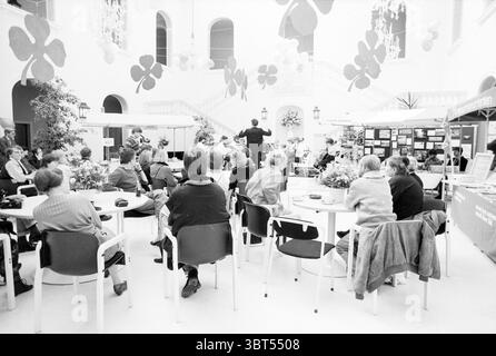 Bloemendaal progressive town hall Bloemendaal Municipality of Bloemendaal Bloemendaal, Whizgle News, Dutch Desk, The Netherlands, 1950 - 2000 on 18-03-1989. These topics appear in the image. In a spacious indoor setting, the atmosphere is lively and communal. The scene features a variety of people sitting at round tables, mostly dressed in comfortable, casual attire, indicating an event or gathering. Some individuals are engaged in conversation, while others are attentively watching a performance occurring in the foreground. Color plays a significant role in the ambiance, with bright, light to Stock Photo