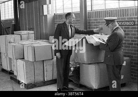 Customs inspection in the customs warehouse in Beverwijk. E. Mooij on the left and A. Fraterman on the right. Beverwijk The Netherlands, Whizgle News, Dutch Desk, The Netherlands, 1950 - 2000 on 30-08-1960. The image shows these topics. The scene depicts a warehouse or storage area with a backdrop of large windows allowing soft, natural light to filter in, casting gentle shadows across the floor. The setting is characterized by industrial elements, including brick walls and concrete flooring, giving it a sturdy, utilitarian feel. In the foreground, two individuals interact amid stacks of cardb Stock Photo