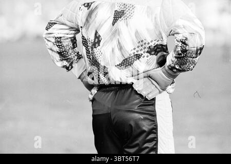 Goalkeeper RCH Heemstede The Netherlands, Whizgle News, Dutch Desk, The Netherlands, 1950 - 2000 on 26-08-1991. These are the elements in the image. The scene captures a figure standing with hands placed firmly on their hips, exuding confidence and focus. The individual is dressed in a vibrant outer garment featuring a bold, abstract pattern, characterized by swirling shapes and contrasting colors of red, blue, and yellow against a predominantly black background. The distinctiveness of the design draws attention, making it a striking element of the scene. Beneath the bright jacket, the person Stock Photo