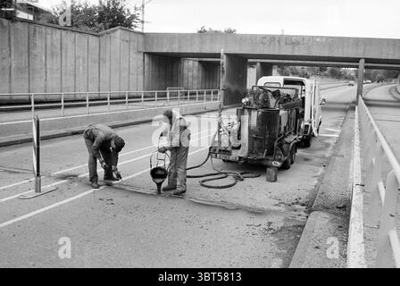 Works viaduct Santpoort-Noord Driehuis Works Driehuis, Whizgle News, Dutch Desk, The Netherlands, 1950 - 2000 on 04-09-1986. These topics are shown in the image. The scene captures a moment of construction work on an urban road, characterized by a monochromatic palette that emphasizes the gritty, industrial atmosphere. Two individuals, clad in protective work attire, are actively engaged in tasks related to road maintenance. One worker is kneeling near the pavement, meticulously pouring a dark mixture from a bucket, while the other stands nearby, overseeing the process and holding equipment. A Stock Photo