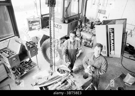 Owner of Derhx Davidson company Velserbroek, Whizgle News, Dutch Desk, The Netherlands, 1950 - 2000 on 12-04-1993. These are the elements in the image. In a busy workshop, two individuals stand prominently in the foreground, both dressed in work overalls that hint at a manual labor environment. They display expressions of concentration and pride, with one man slightly leaning forward, suggesting engagement in the activity at hand. Surrounding them are various tools and mechanical parts scattered across a workbench, showcasing an air of industriousness. A metal toolbox, filled with assorted too Stock Photo