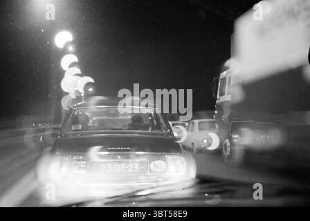File, Whizgle News, Dutch Desk, The Netherlands, 1950 - 2000 on 12-1978. These are the elements in the image. The scene captures a nighttime urban setting viewed through a windshield, with a focus on the blurred figures of cars ahead. The leading vehicle, characterized by its vintage design, displays a dark color that contrasts against the brighter reflective lights around it. Brilliant orbs of light from street lamps and vehicle headlights punctuate the foreground, creating a dreamy, ethereal quality. These lights refract softly, resulting in a bokeh effect that enhances the scene's atmospher Stock Photo