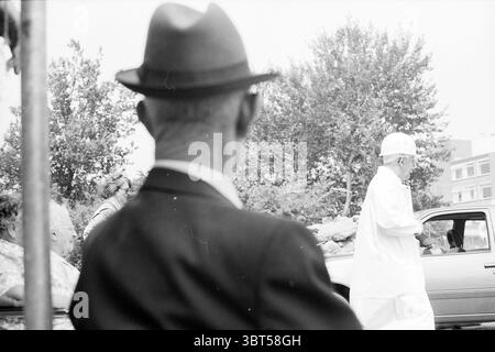 First post Lisse, Whizgle News, Dutch Desk, The Netherlands, 1950 - 2000 on 04-07-1991. The image includes these topics. In a scene characterized by a nostalgic black-and-white aesthetic, an elderly man is positioned in the foreground, partially obscured by a dark, wide-brimmed hat that casts a slight shadow over his face. His attire is formal, featuring a dark suit that adds to the sense of an older era. The background reveals another figure—an individual dressed in a light, flowing garment, likely cultural or traditional in style, suggesting a sense of significance. This figure is in motion, Stock Photo