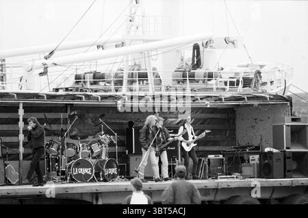 Pop concert Loggerstraat IJmuiden IJmuiden Loggerstraat Nederland, Whizgle News, Dutch Desk, The Netherlands, 1950 - 2000 on 16-07-1993. These are the elements in the image. The scene captures a lively outdoor performance on a boat or a large structure elevated above its surroundings. The setting is dynamic, suggesting a maritime atmosphere with a hint of adventure. In the foreground, a small crowd gathers, their figures slightly out of focus, showing interest in the performance. The people appear engaged, with some perhaps swaying or tapping their feet to the music. They are dressed in casual Stock Photo