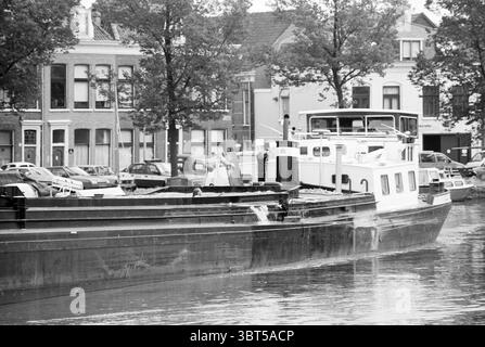 Swimming in a cargo boat Spaarne, Whizgle News, Dutch Desk, The Netherlands, 1950 - 2000 on 14-07-1992. These topics are shown in the image. The scene features a large, industrial vessel gliding through calm waters, which reflect the surrounding environment. The boat, predominantly black and white, contrasts with the muted tones of the water. Its structure is robust, with a flat deck and a cabin featuring large windows. In the background, several buildings stand together, showcasing a mixed architectural style. They include traditional facades with brick and stucco elements, primarily in shade Stock Photo