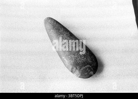 Ancient stone hand axe, Whizgle News, Dutch Desk, The Netherlands, 1950 - 2000 on 27-03-1991. The image contains these topics. The scene features a smooth, elongated stone positioned prominently against a textured, light-colored surface. The stone has a muted gray tone, with subtle variations in shading that highlight its natural contours and surface imperfections. The grainy texture of the stone contrasts with the smoothness of the background, which appears to be a soft, muted hue that enhances the stone’s features. Lighting is soft but directional, casting gentle shadows that emphasize the s Stock Photo