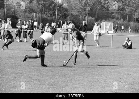 Strawberries - Apeldoorn Hockey, Whizgle News, Dutch Desk, The Netherlands, 1950 - 2000 on 25-09-1983. These are the elements in the image. The scene depicts an energetic field hockey match taking place on a bright, sunny day. Two players are engaged in a competitive moment; one, in dark shorts and a lighter top, is skillfully maneuvering the ball towards the opposing player. The other, wearing a dark shirt and white shorts, is poised to intercept, focusing intently on the ball. In the background, a crowd of spectators is gathered along the sidelines, dressed in casual clothing. Some are sitti Stock Photo
