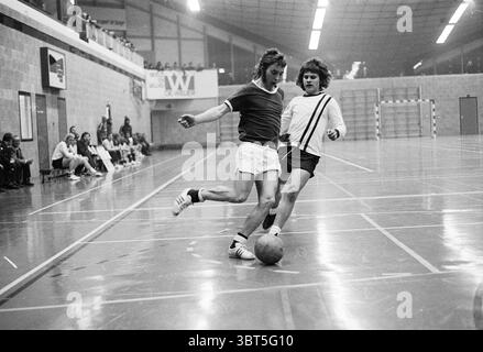 Indoor Football Championship Football, Whizgle News, Dutch Desk, The Netherlands, 1950 - 2000 on 18-04-1973. These are the elements in the image. The scene captures an intense moment in an indoor sports setting, predominantly featuring two players engaged in a dynamic game of soccer. The athletes are dressed in distinct uniforms: one wears a short-sleeved, dark top paired with light shorts, while the other is clad in a lighter shirt and dark shorts, showcasing a stark contrast in colors. The floor is polished and smooth, reflecting the overhead lights, which illuminate the space with a bright, Stock Photo