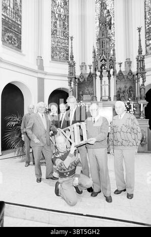 Group of handymen in Laurentius Church Heemskerk, Whizgle News, Dutch Desk, The Netherlands, 1950 - 2000 on 02-12-1990. The image contains these topics. The scene captures a group of individuals gathered inside a grand hall, characterized by ornate architectural details and a sense of reverence. In the background, an intricate stained glass window illuminates the space with soft, diffused light, casting subtle colorful patterns on the floor. The group features several older men, primarily dressed in tailored suits and cardigans, exhibiting a sense of camaraderie. They stand together in a semi- Stock Photo