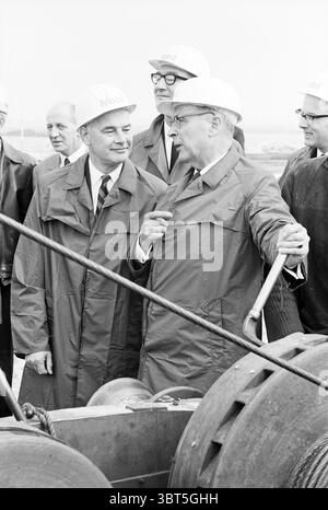 Driving the first pile., Whizgle News, Dutch Desk, The Netherlands, 1950 - 2000. The image contains these topics. In a subdued, monochromatic setting, a group of men is gathered, all wearing similar hard hats, suggesting a formal or industrial occasion. They are dressed in long coats, often associated with outdoor or professional environments. The men stand close together, engaged in conversation, with a sense of camaraderie or collaboration. The foreground features machinery, possibly indicative of construction or technological work, adding an industrial feel to the scene. The composition app Stock Photo