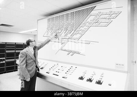 MSD warehouse + computer Haarlem Buildings, Whizgle News, Dutch Desk, The Netherlands, 1950 - 2000 on 07-10-1977. The image includes these topics. The scene is set in a large, functional room with a modern yet industrial ambiance. A man stands to the left, dressed in a formal, grey suit with a distinct, patterned tie, which adds a touch of sophistication to the environment. He is pointing towards a large technical diagram mounted on the wall, which outlines a complex layout, possibly related to machinery or a computing system. The diagram is predominantly white with various lines and schematic Stock Photo
