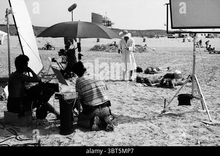Avro TV recordings beach IJmuiden IJmuiden The Netherlands, Whizgle News, Dutch Desk, The Netherlands, 1950 - 2000 on 18-08-1995. The image shows these topics. The scene unfolds on a sandy beach, vibrant yet subdued in a monochromatic palette. The sun casts a bright, harsh light, creating stark contrasts between the shadows and highlights. In the foreground, a couple of individuals kneel on the sand, their postures indicating focus as they operate equipment seemingly for a film or photography setup. One person is dressed in casual beach attire, while the other dons a structured shirt with a di Stock Photo
