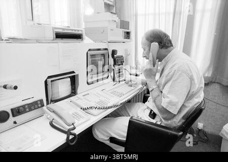 Operator in a control room, Whizgle News, Dutch Desk, The Netherlands, 1950 - 2000 on 05-09-1989. These topics are shown in the image. In a well-lit room characterized by soft, diffused lighting filtering through sheer curtains, a man is seated at a large white desk cluttered with various objects. He wears a light-colored, casual shirt and is engaged in a conversation on a rotary telephone held to one ear while manipulating equipment on the desk. The desk features two computer monitors, displaying old-style screens with visible geometric patterns. One monitor is larger, positioned to his right Stock Photo