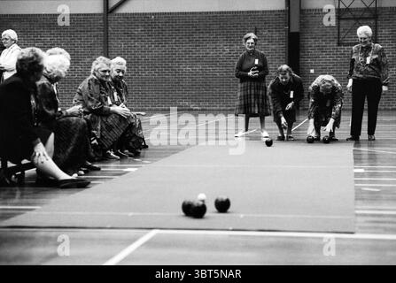Elderly race ball sports hall Zeewijk games and toys IJmuiden Planetenweg The Netherlands, Whizgle News, Dutch Desk, The Netherlands, 1950 - 2000 on 26-02-1990. The image includes these topics. The scene is set indoors in a spacious, well-lit hall with a polished wooden floor that reflects the ambient light. The walls are a muted brick red, providing warmth to the setting. In the foreground, a long, narrow green mat runs the length of the composition, serving as the playing area. Several elderly women are engaged in a lawn bowling activity. Two of them are focused on rolling balls down the mat Stock Photo