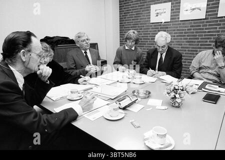 Zwanenburg Village Council meeting Zwanenburg, Whizgle News, Dutch Desk, The Netherlands, 1950 - 2000 on 12-04-1995. These are the topics in the image. A group of six individuals is seated around a rectangular table, engaged in a discussion. The setting has a warm and inviting atmosphere, highlighted by soft ambient lighting that illuminates the room. The walls are adorned with light-colored brickwork, giving the environment a cozy, professional feel. The table is cluttered with various documents, notebooks, and pens, indicating a working meeting. Small, elegant cups and saucers are placed in Stock Photo