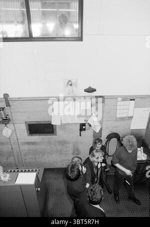 Last Typhoon, Whizgle News, Dutch Desk, The Netherlands, 1950 - 2000 on 13-02-1992. These topics are shown in the image. In a spacious room characterized by a minimalist industrial aesthetic, the atmosphere is subdued and somewhat chaotic, reflecting a busy environment. The floor is made of dark, textured material, while the walls are a mix of concrete and plaster, giving a utilitarian feel to the space. Light filters in from a high window, casting soft shadows and creating areas of contrast. A small group of people congregates in the lower part of the scene. Their styles vary, with some dress Stock Photo