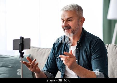 Attractive mature man broadcasting from home, using smartphone on tripod Stock Photo