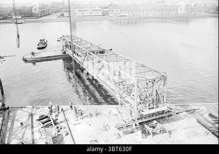 An iron construction above the water., Whizgle News, Dutch Desk, The Netherlands, 1950 - 2000. The image includes these topics. The scene captures a dynamic construction site along a riverbank. In the foreground, construction workers are engaged in various activities, indicating a collaborative effort. They are dressed in typical work attire, such as helmets and rugged clothing, reflecting an industrial environment. The main focus is on a large steel structure being assembled, its intricate framework stark against the calm waters of the river. The steel beams, painted in a somewhat muted tone, Stock Photo