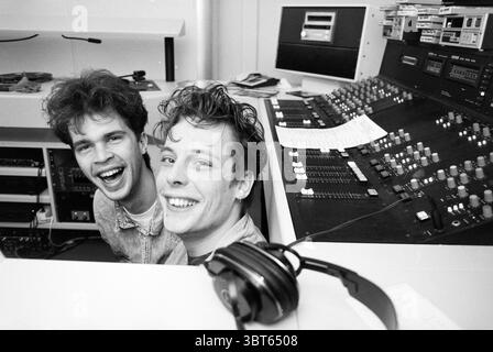 2 DJs at Radio 10 A'dam Amsterdam The Netherlands, Whizgle News, Dutch Desk, The Netherlands, 1950 - 2000 on 19-02-1991. These are the topics in the image. In a lively recording studio, two young men share a joyful moment, their faces bright with smiles and infectious laughter. Both have tousled hair, with the texture appearing slightly curly, and they wear casual denim jackets that give off a relaxed vibe. The setting feels energetic and creative, with a backdrop filled with an array of audio equipment. The large mixing console in front of them is a focal point, adorned with numerous colorful Stock Photo