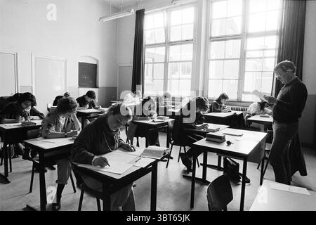 Final Greek exam gymnasium Velsen Zuid Exam Velsen-Zuid, Whizgle News, Dutch Desk, The Netherlands, 1950 - 2000 on 26-04-1985. These are the topics in the image. The scene is set in a classroom filled with natural light streaming in through large windows, illuminating the space with a soft glow. The walls are painted in a light hue, enhancing the airy feel of the room. Desks are arranged in rows, each occupied by students who are bent over their work, absorbed in their tasks. Their expressions vary; some look focused, while others appear deep in thought. Each student is equipped with writing m Stock Photo