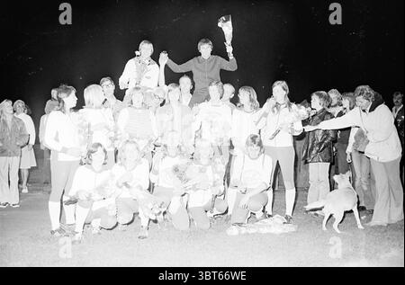 Softball Terrasvogels - Bloemendaal (Terrasvogels champion) Softball, Whizgle News, Dutch Desk, The Netherlands, 1950 - 2000 on 06-09-1975. The image includes these topics. The scene is set in a lively nighttime environment, illuminated by soft artificial lighting that casts a warm glow over the group. In the foreground, a diverse group of individuals, mostly dressed in sports attire, stands in a dynamic pose. Some are kneeling while others are proudly holding bouquets, suggesting a celebratory event, possibly a sports victory. The individuals express a mix of emotions ranging from joy to exci Stock Photo