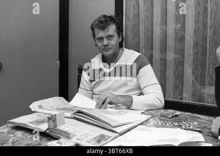 Henny van Egdom former Elinkwijk football player Football people and trainers, Whizgle News, Dutch Desk, The Netherlands, 1950 - 2000 on 28-11-1983. The image includes these topics. The scene depicts a man seated at a table, exuding a sense of calm and introspection. He is dressed in a striped sweater featuring shades of white and grey, with a casual, relaxed demeanor. His hair is slightly tousled, suggesting a comfortable yet unrefined look. The table in front of him is cluttered with various items, including open magazines, newspapers, and a few scattered papers, indicating a moment of study Stock Photo