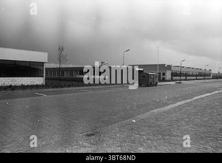 Exterior sheltered workshop 'De Tunnel' Velsen-Noord Cruquiusweg, Whizgle News, Dutch Desk, The Netherlands, 1950 - 2000 on 11-04-1972. These topics appear in the image. The scene depicts a somber urban landscape under overcast skies, casting a muted light across the surroundings. On one side, a row of low-rise buildings features a modern design, characterized by clean lines and large windows, hinting at their commercial or industrial purpose. The building on the left has a glass façade, which reflects the cloudy sky, while the structure beside it has a brick facade that contributes to a more Stock Photo