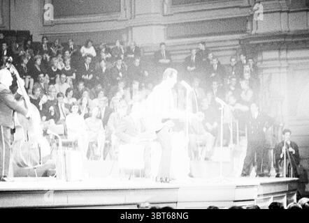 Trini Lopez in Amsterdam Concertgebouw Music, Whizgle News, Dutch Desk, The Netherlands, 1950 - 2000 on 13-10-1963. These are the topics in the image. The scene captures a concert or performance in a grand hall filled with a captivated audience. The lighting is soft and focused primarily on the performers, creating a stark contrast with the dimly lit seating area where the crowd sits. On stage, a male musician is front and center, holding a guitar, his figure slightly blurred, emphasizing movement. He appears engaged with the audience, possibly singing or playing passionately. Surrounding him Stock Photo