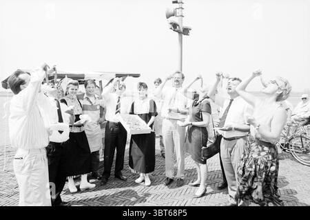 Tasting herring College Velsen, Whizgle News, Dutch Desk, The Netherlands, 1950 - 2000 on 21-05-1992. These are the topics in the image. In the scene, a diverse group of individuals is gathered near a street vendor, likely celebrating a joyful occasion. The people are dressed in a mix of casual and semi-formal attire, creating a lively atmosphere. Some are wearing light-colored garments, while others sport darker hues, providing a contrast against the bright, clear sky above. The expressions on their faces reveal excitement and camaraderie, with many smiling and laughing, suggesting a playful Stock Photo