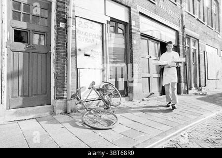 Bakery in demolished buildings, Whizgle News, Dutch Desk, The Netherlands, 1950 - 2000 on 24-08-1994. These are the elements in the image. In a gritty urban setting, a narrow street features a mix of brick and wooden buildings, some of which show signs of age and neglect. The foreground is dominated by an overturned bicycle, its wheel distorted, suggesting a past mishap. Nearby, a figure dressed in a white shirt and apron walks slightly away from the camera, holding a flat rectangular object, possibly a pizza or a tray of food, balancing it carefully. The mood feels somber yet lively, with a h Stock Photo