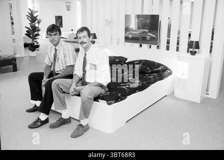 New bed store Heemstede shops and kiosks Heemstede The Netherlands, Whizgle News, Dutch Desk, The Netherlands, 1950 - 2000 on 07-08-1989. These topics are shown in the image. The scene features two men seated on a modern, stylish bed that showcases a sleek design. The bed has a white frame with an inviting mattress adorned with a patterned black and white bedspread, which features abstract designs. Both men display distinctive styles. One has dark, wavy hair, wearing a striped button-down shirt complemented by dark pants and polished shoes. The other has a more classic look with a mustache, dr Stock Photo