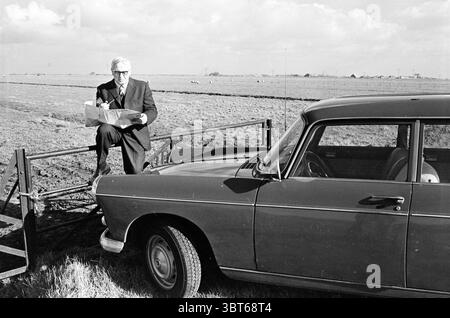 man with pipe road map and Peugeot 404 in meadow between ponies., Whizgle News, Dutch Desk, The Netherlands, 1950 - 2000. The image shows these topics. In a vast, open landscape, a man dressed in a formal dark suit and light-colored shirt sits on the gate, casually holding a paper container of food. He has neatly styled light hair and wears glasses, giving off an air of sophistication. The vehicle beside him is a classic black car, its shiny surface catching the light under a partly cloudy sky, which features billowing white clouds against a backdrop of pale blue. The foreground is dominated b Stock Photo