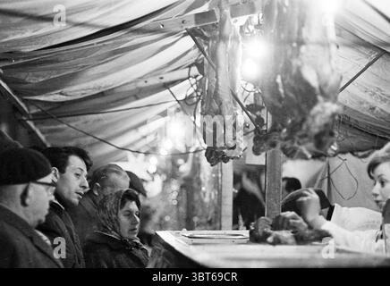 Christmas market at Butter Market Christmas Christmas Christmas decorations Christmas decorations Market Haarlem Botermarkt The Netherlands, Whizgle News, Dutch Desk, The Netherlands, 1950 - 2000 on 23-12-1963. These are the topics in the image. The scene unfolds under a dimly lit canopy, where muted, warm lights cast a soft glow, illuminating the gathering of people engaged in conversation. A rustic stall acts as the focal point, displaying various cuts of meat hanging from hooks, their textures contrived in shades of deep brown and red. To one side, a group of individuals stands closely, the Stock Photo