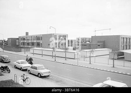 Beverwijk School Community Schools, Whizgle News, Dutch Desk, The Netherlands, 1950 - 2000 on 23-10-1970. These are the elements in the image. The scene depicts a modest urban area dominated by a school building. The architecture features rectangular structures, characterized by brick exteriors in muted tones of gray and brown. Large windows punctuate the façade, reflecting a cloudy sky overhead. A few trees with sparse foliage line the walkway, adding a touch of softness amidst the rigid architecture. In the foreground, a street runs parallel to the school, with a few parked vehicles. Among t Stock Photo