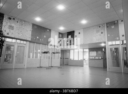 Wall painting Polder House, Whizgle News, Dutch Desk, The Netherlands, 1950 - 2000 on 21-11-1991. These topics are shown in the image. The scene depicts a spacious interior area characterized by high ceilings and a broad floor made of light-colored tiles. The walls are adorned with a series of abstract artworks in varying muted tones—reds, blues, yellows, and grays. These artworks create a colorful mosaic effect that brings a sense of vibrancy to the otherwise neutral environment. To one side, a set of glass double doors leads to another area, allowing natural light to pour in, illuminating th Stock Photo