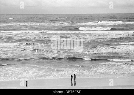 Strong wind at sea and on the beach., Whizgle News, Dutch Desk, The Netherlands, 1950 - 2000. The image contains these topics. The scene captures two figures standing at the edge of a vast, tumultuous ocean, their silhouettes sharply defined against the shoreline. The beach stretches out in soft, pale sands, contrasting with the darker, swirling waters of the sea. The waves roll in with a rhythmic energy, crowned with frothy white crests that contrast against the deep blue-gray of the ocean. Overhead, the sky is overcast with heavy clouds, lending a subdued and moody atmosphere to the scene. T Stock Photo