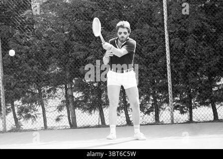 Tennis. Men's doubles and mixed doubles, Whizgle News, Dutch Desk, The Netherlands, 1950 - 2000 in 1981. These are the topics in the image. In the scene, a man is poised on a tennis court, captured mid-swing with a racket in hand. He sports a dark polo shirt and white shorts that highlight his active stance. His sunglasses reflect a bright, sunny day, adding an air of cool confidence. The surrounding area is enclosed by a chain-link fence, and just beyond it, there are dense patches of greenery, indicating a well-maintained outdoor setting. The court's surface is a muted shade, contrasting wit Stock Photo
