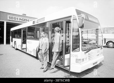 New Bus NZH, Whizgle News, Dutch Desk, The Netherlands, 1950 - 2000 on 26-07-1990. The image shows these topics. In the scene, a large, modern bus dominates the foreground, showcasing a clean, white exterior adorned with minimalistic branding. The bus's doors are open, revealing a spacious interior. A few individuals are visible; one man stands confidently at the base of the bus steps, wearing a suit and exuding an air of professionalism, while another figure, slightly obscured, appears to be stepping off. The setting conveys a transportation hub, likely a depot or garage, with a hint of indus Stock Photo