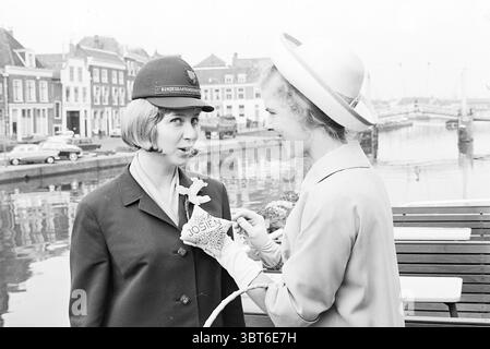 Haarlem. flower girls on ship 'city of Essen'. Flowers and flower girls Haarlem Spaarne The Netherlands, Whizgle News, Dutch Desk, The Netherlands, 1950 - 2000 on 09-04-1965. The image includes these topics. In a serene urban setting, two women engage in a lively moment by a calm waterway. One woman wears a dark uniform with a distinctive hat, projecting an air of authority and professionalism. Her hairstyle is neatly styled and frames her face, highlighting her expression of intrigue or surprise as she interacts with the other. The second woman, dressed in a light-toned garment that exudes el Stock Photo