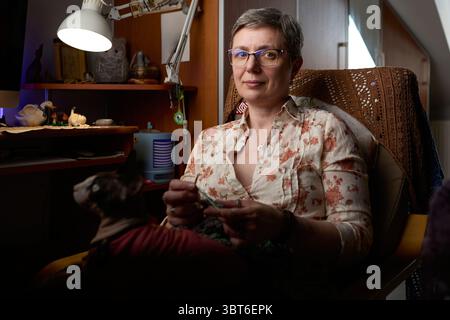 A confident mature woman sitting in a cozy home environment, crocheting a handmade piece. A Sphynx cat in a red sweater is sitting on her lap. Stock Photo