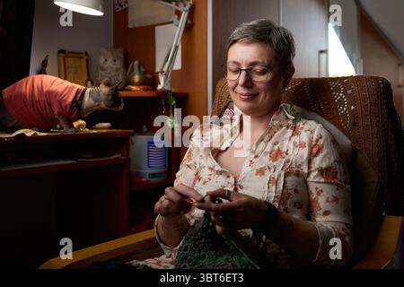 A confident mature woman sitting in a cozy home environment, focused on crocheting a handmade piece. In the background, a Sphynx cat in a red sweater Stock Photo