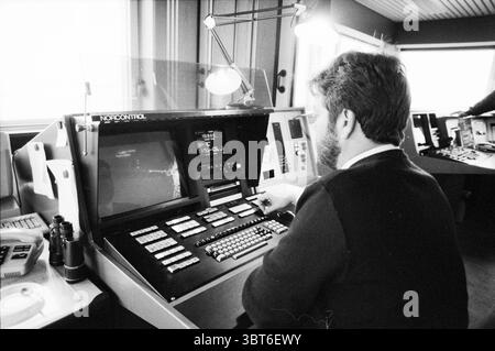 Pilotage employee behind control computer IJmuiden The Netherlands, Whizgle News, Dutch Desk, The Netherlands, 1950 - 2000 on 07-12-1989. These are the topics in the image. The scene captures a focused moment in a control room, set in a sleek, modern environment. In the foreground, a man is seated at a large, intricate console filled with an array of buttons, switches, and screens. His attention is drawn to one particular monitor displaying data, illuminated by a soft glow, which contrasts with the otherwise muted tones of the surroundings. The man has a neat beard and is dressed in a dark swe Stock Photo