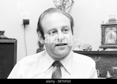 Portrait of a man about 50 years old in the living room, Whizgle News, Dutch Desk, The Netherlands, 1950 - 2000 on 15-06-1979. These are the elements in the image. The scene features a man seated in a cozy indoor setting. His expression is calm and engaged, suggesting a relaxed demeanor. He has dark, slicked-back hair and is dressed in a light-colored shirt with a patterned tie that adds a touch of sophistication to his appearance. Behind him, the background is softly blurred, drawing attention to his face. The setting contains elements of a home or office, with a dark wooden cabinet visible, Stock Photo