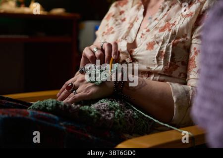 Close-up of a woman crocheting a handmade green and gray floral pattern using a crochet hook. She is seated indoors, wearing a floral blouse Stock Photo