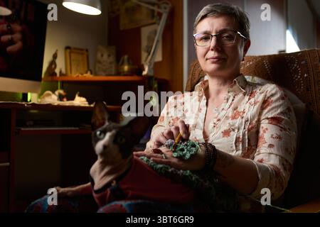 A confident mature woman sitting in a cozy home environment, crocheting a handmade piece. A Sphynx cat in a red sweater is sitting on her lap. The war Stock Photo