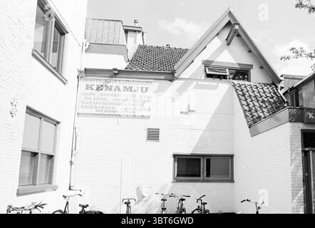 Kenamju exterior and Jazz Ballet, Whizgle News, Dutch Desk, The Netherlands, 1950 - 2000 on 03-05-1994. These topics are shown in the image. The scene depicts a charming urban alleyway, showcasing a blend of architectural styles. The primary structure is a white building with a textured, plaster-like facade that reflects sunlight. Its gabled roof features distinct tiles, lending a traditional yet quaint appearance to the architecture. Several bicycles are lined up neatly against the wall in the foreground, their frames painted in muted tones, suggesting a casual, everyday atmosphere. Each bike Stock Photo