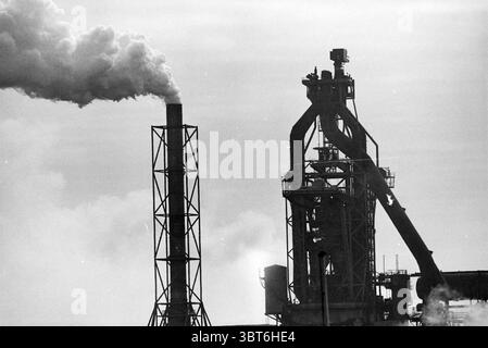 Blast furnaces various, Whizgle News, Dutch Desk, The Netherlands, 1950 - 2000 on 18-03-1992. These topics appear in the image. The scene presents a stark industrial landscape characterized by towering structures and billowing smoke. Dominating the composition is a tall chimney, its silhouette bold against a pale sky, from which dense, white smoke ascends in a tapered plume. The smoke contrasts sharply with the lighter background, creating a dramatic effect. To the right, an imposing industrial facility is visible, with various pipes and frameworks intertwining in a complex, almost chaotic man Stock Photo