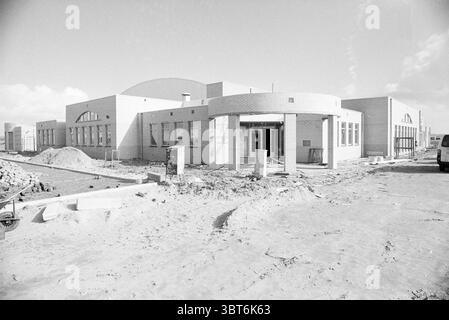 Ext. Polder house V'broek Exterior Velserbroek, Whizgle News, Dutch Desk, The Netherlands, 1950 - 2000 on 09-03-1990. These topics appear in the image. The composition showcases a newly constructed building with a modern architectural design. The structure features a combination of smooth and textured surfaces, predominantly in shades of light gray and beige, contributing to a clean and contemporary look. Surrounding the building is a rough, unfinished landscape characterized by patches of dirt and gravel, indicating ongoing construction. Some materials, such as bricks and stones, are scattere Stock Photo
