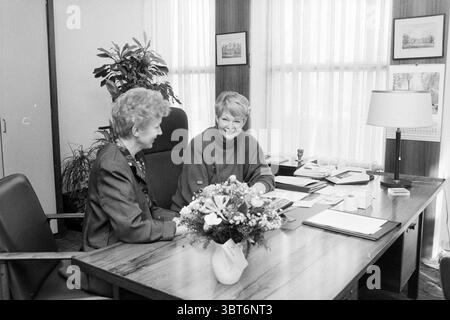 1st working day Mayor Van Dok IJmuiden IJmuiden The Netherlands, Whizgle News, Dutch Desk, The Netherlands, 1950 - 2000 on 05-02-1990. These are the topics in the image. In a well-lit office setting, two women are seated at a large wooden desk, engaging in a friendly conversation. The desk is organized with various documents and a small lamp, casting soft light across the surface. A bouquet of flowers in a vase adds a pop of color, contrasting beautifully with the monochromatic aesthetic of the scene. The room is decorated with potted plants, gently enhancing the atmosphere with touches of gre Stock Photo