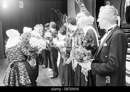 VVV Zandvoort gives flowers to players of Madame Arthur Musical Zandvoort, Whizgle News, Dutch Desk, The Netherlands, 1950 - 2000 on 18-12-1985. These topics are shown in the image. The scene is set on a stage, likely in a theater, illuminated with soft yet dramatic lighting that casts gentle shadows across the space. In the foreground, a diverse group of elegantly dressed individuals stands in a line, celebrating a special occasion. The people in the scene express various emotions, ranging from joy to pride. The men are dressed in classic tuxedos, complete with bow ties, while the women wear Stock Photo
